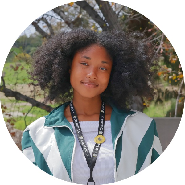 A woman with black hair and a turquoise jacket smiling toward the camera. green foliage in the background.