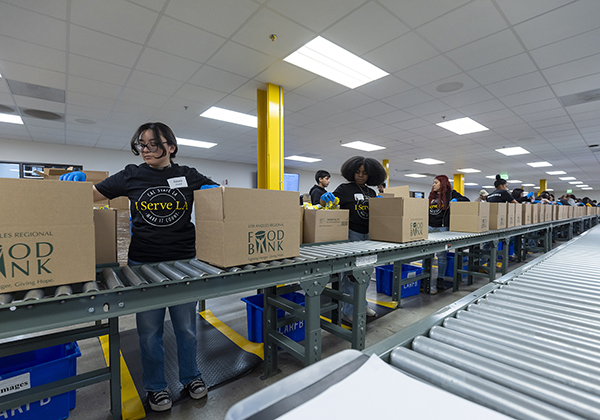 Cardboard boxes travel along a conveyor belt in front of students wearing matching t-shirts.