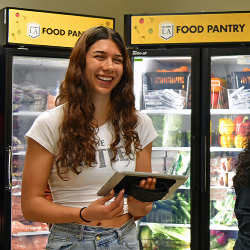 Smiling long-haired person holds a clipboard in front of Cal State LA Food Pantry refrigerators now.