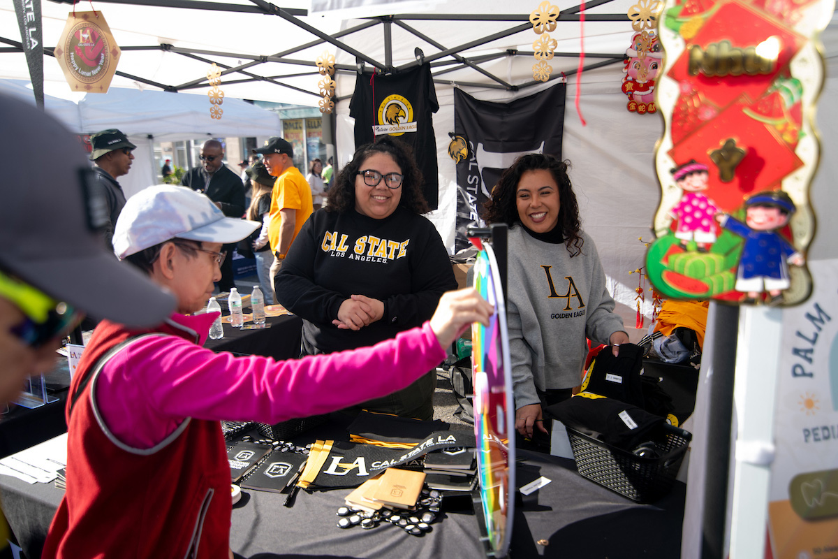 Two women wearing cal state l.a. sweatshirts smiling under a tent as a patron spins a raffle wheel