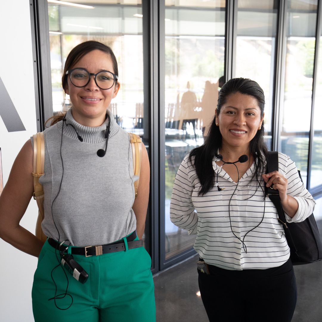 Two women stand indoors, smiling at the camera. Both are wearing headsets with microphones.