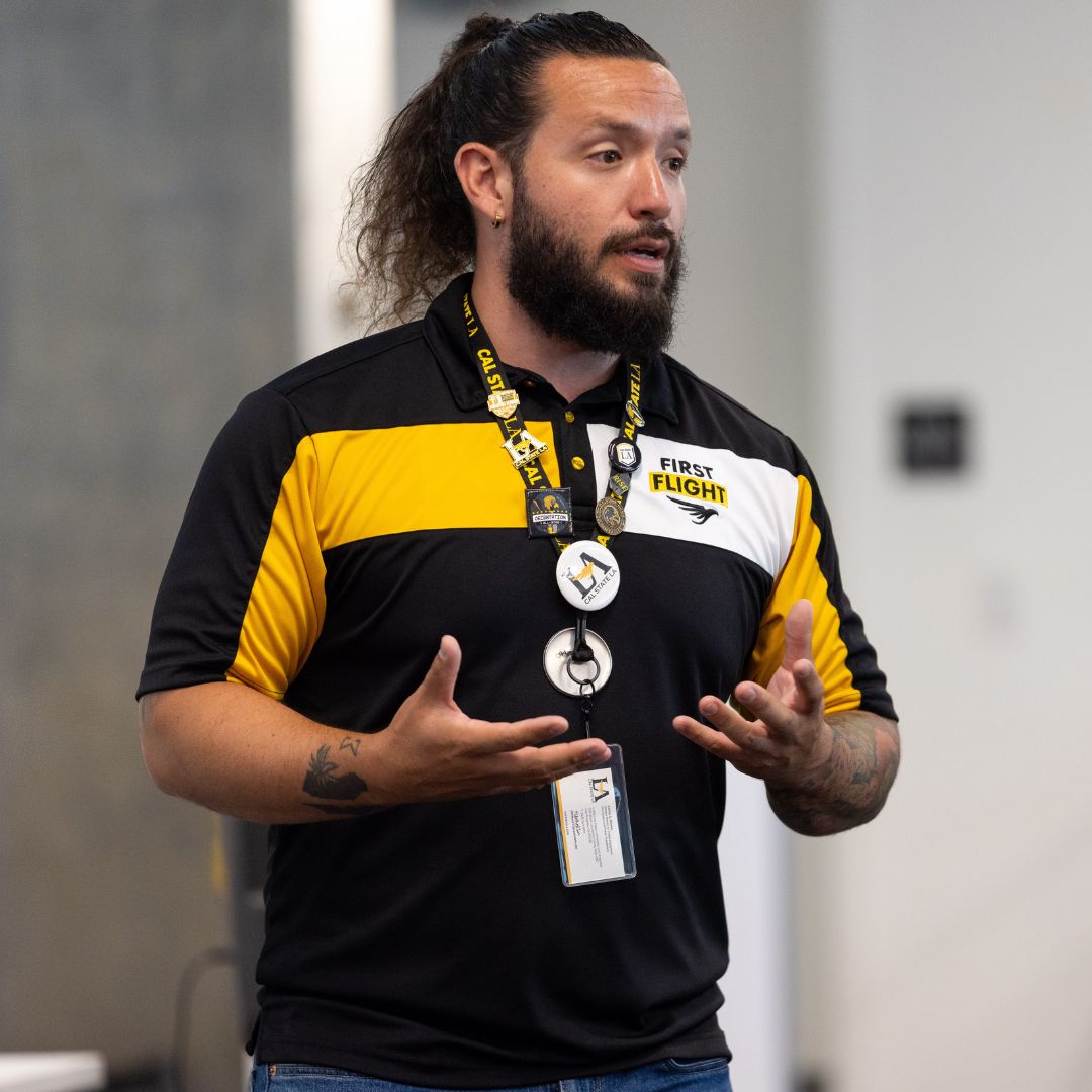 A man wearing a black and yellow shirt stands speaking and gesturing with his hands. 