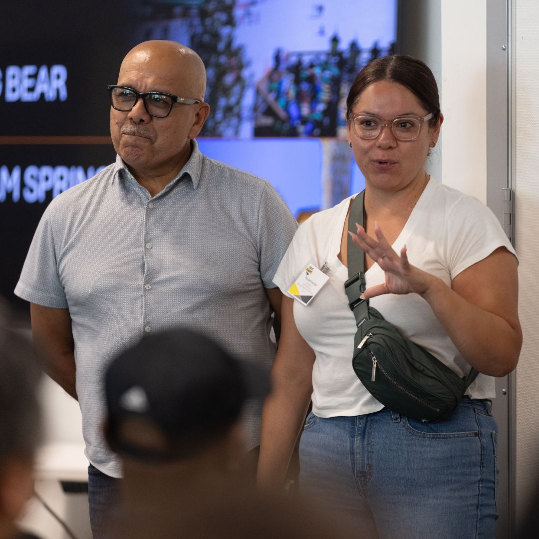 Two adults stand at the front of a room addressing a group. 