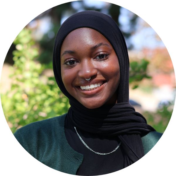 A woman in a hijab smiling towards the camera. There are trees in the background.