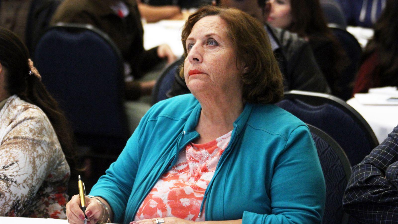 A woman in a teal jacket listens attentively while holding a pen at a conference table