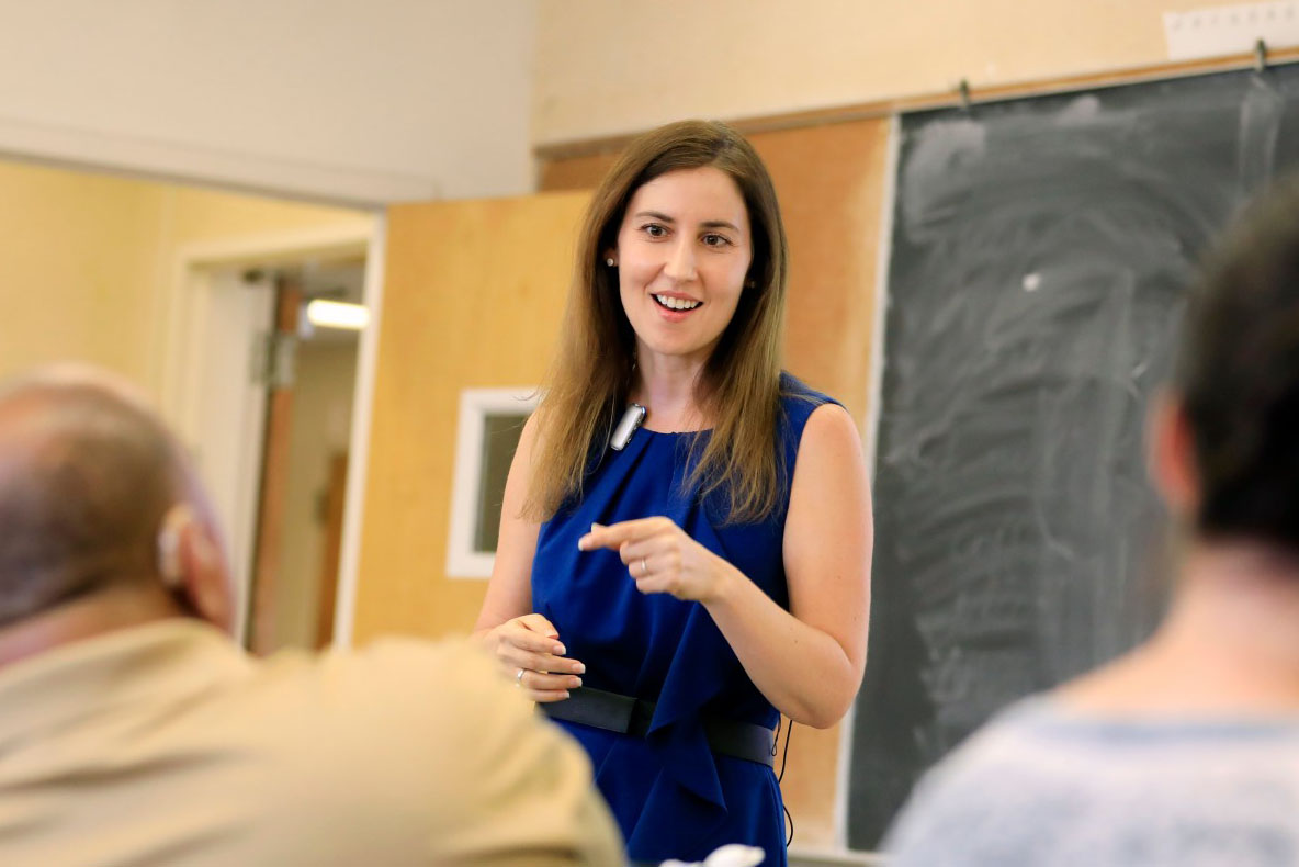 A woman in a blue dress speaks and gestures while teaching in front of a classroom chalkboard