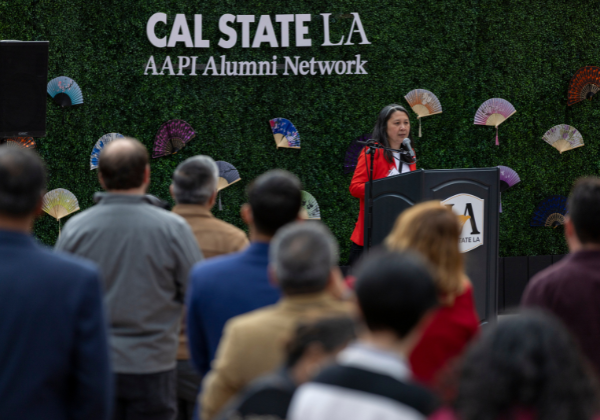 Large group of people listening to a speaker at a podium. Green hedges in the background with sign that says Cal State LA AAPI Alumni Network