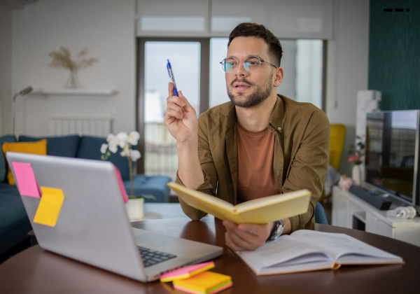 Male student at home sitting at desk with open laptop. He is holding a text book and talking during an online class.