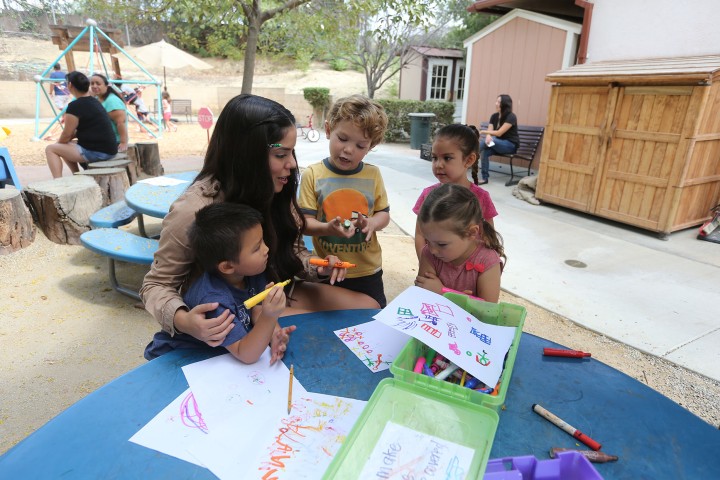 An adult sits with four young children at an outdoor table, drawing with crayons and markers 