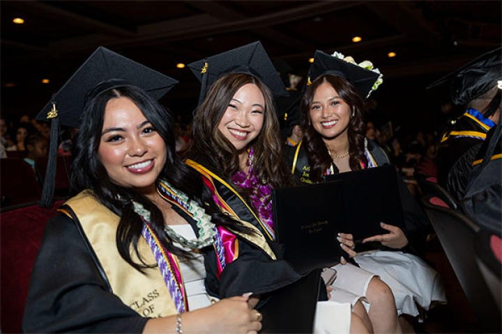 Three graduates in caps and gowns sit smiling in an auditorium during commencement.