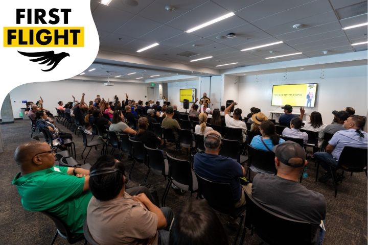 A large group of people sits in a conference-style room. A “First Flight” logo appears in the corner, suggesting this is part of a program or workshop session