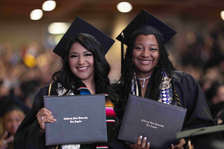 Two graduates wearing their cap and gown smiling and holding up diploma