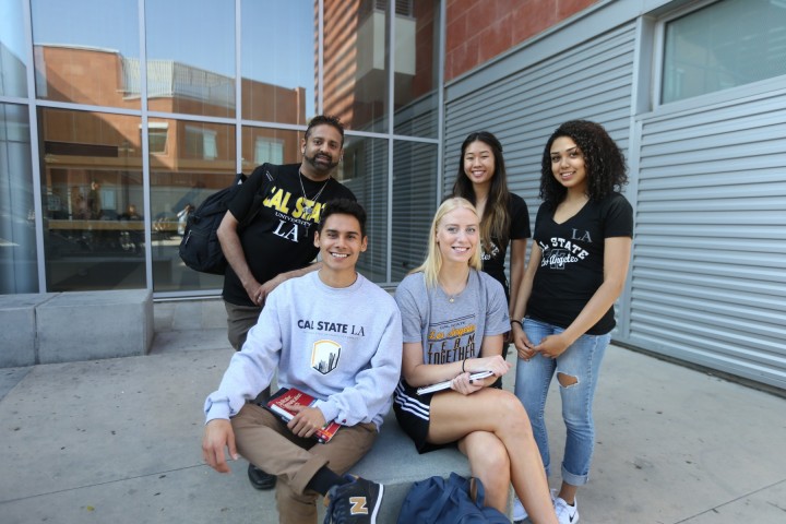 five students standing in front of a building with glass. Two students sitting down and three right behind