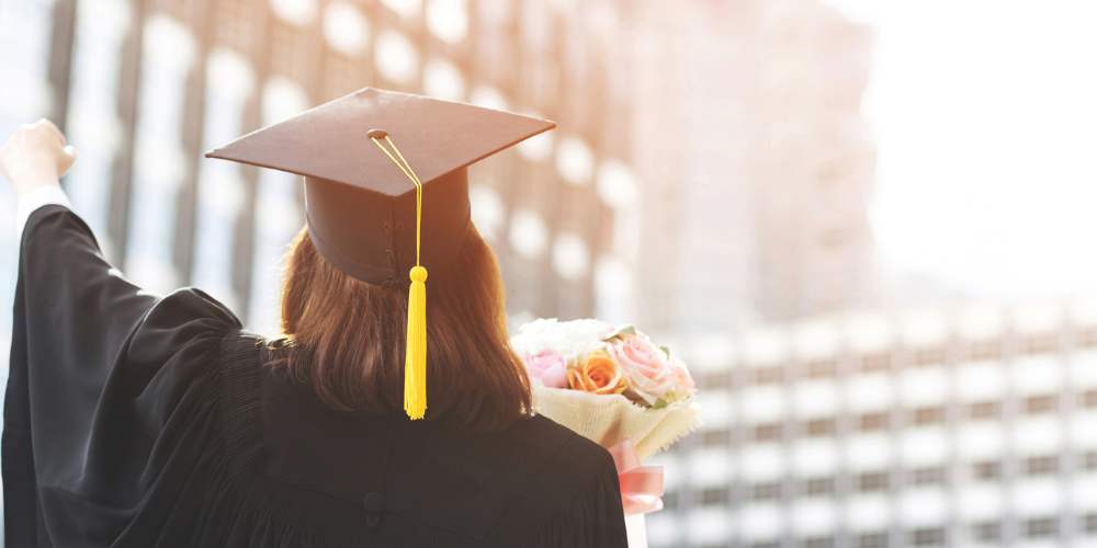 Female graduate wearing black cap and gown, holding flowers with one hand, and raising the other in celebration.