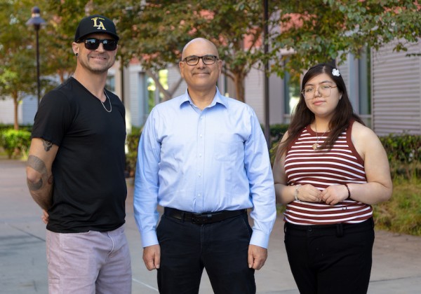 A man in a black shirt and baseball cap stands on the left, Professor Jamil Momand stands in the center wearing a light blue button-down shirt, and a student stands on the right wearing a red-and-white striped sleeveless top.