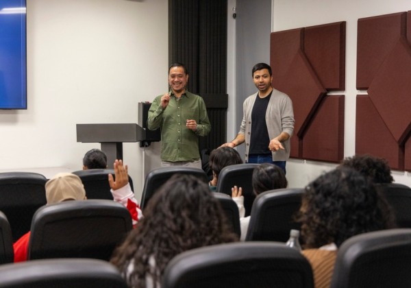 Two presenters stand at the front of a classroom speaking to a seated audience, with students raising their hands during a discussion.