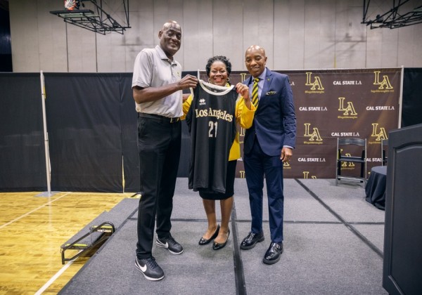 Three individuals stand on a stage holding a Los Angeles jersey during a Cal State LA athletics event.