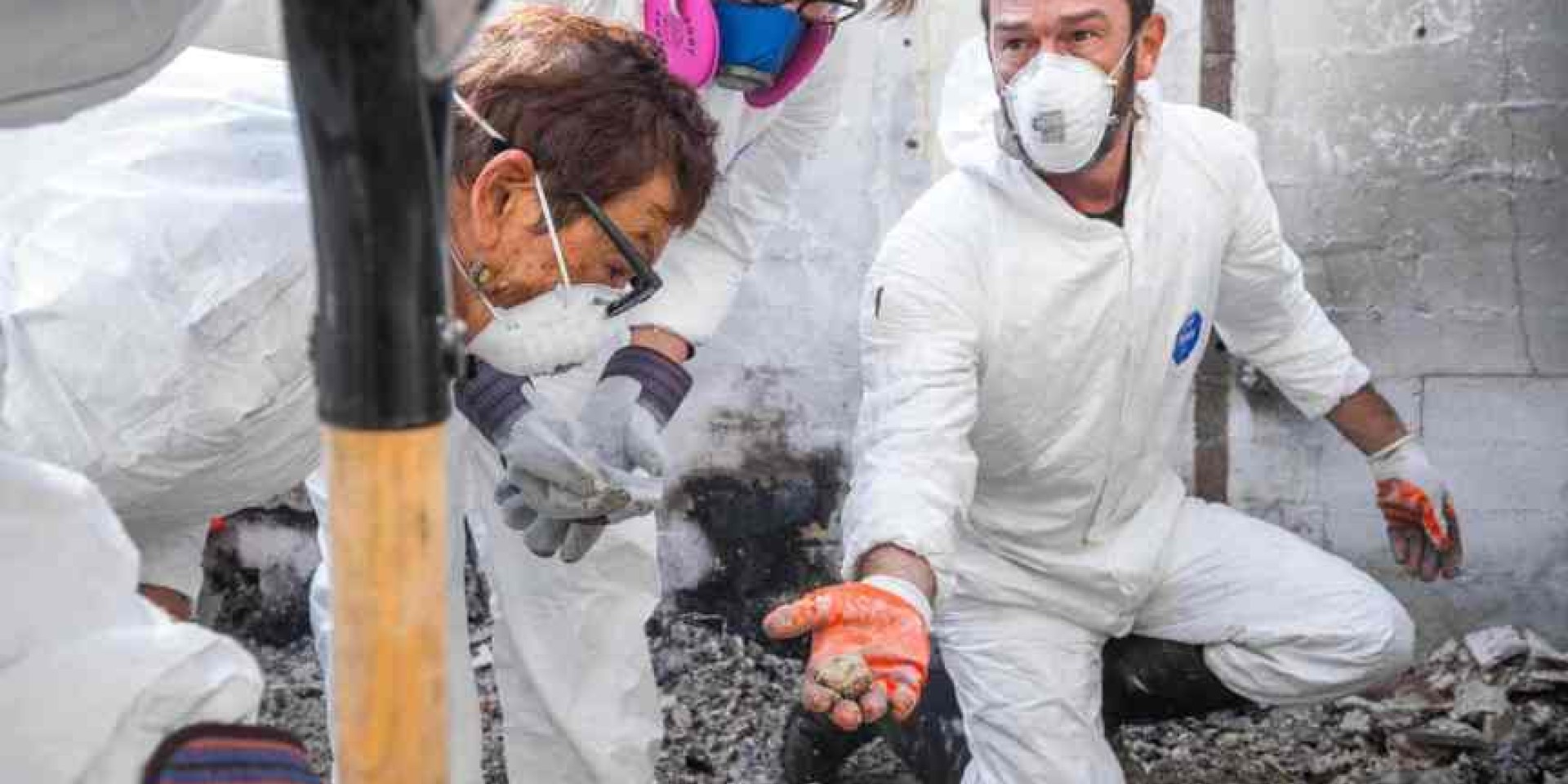 A masked archaeologist kneels in the rubble of a burned home, carefully holding a small, recovered object while colleagues look on.