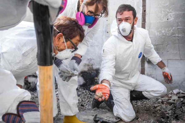 A masked archaeologist kneels in the rubble of a burned home, carefully holding a small, recovered object while colleagues look on.